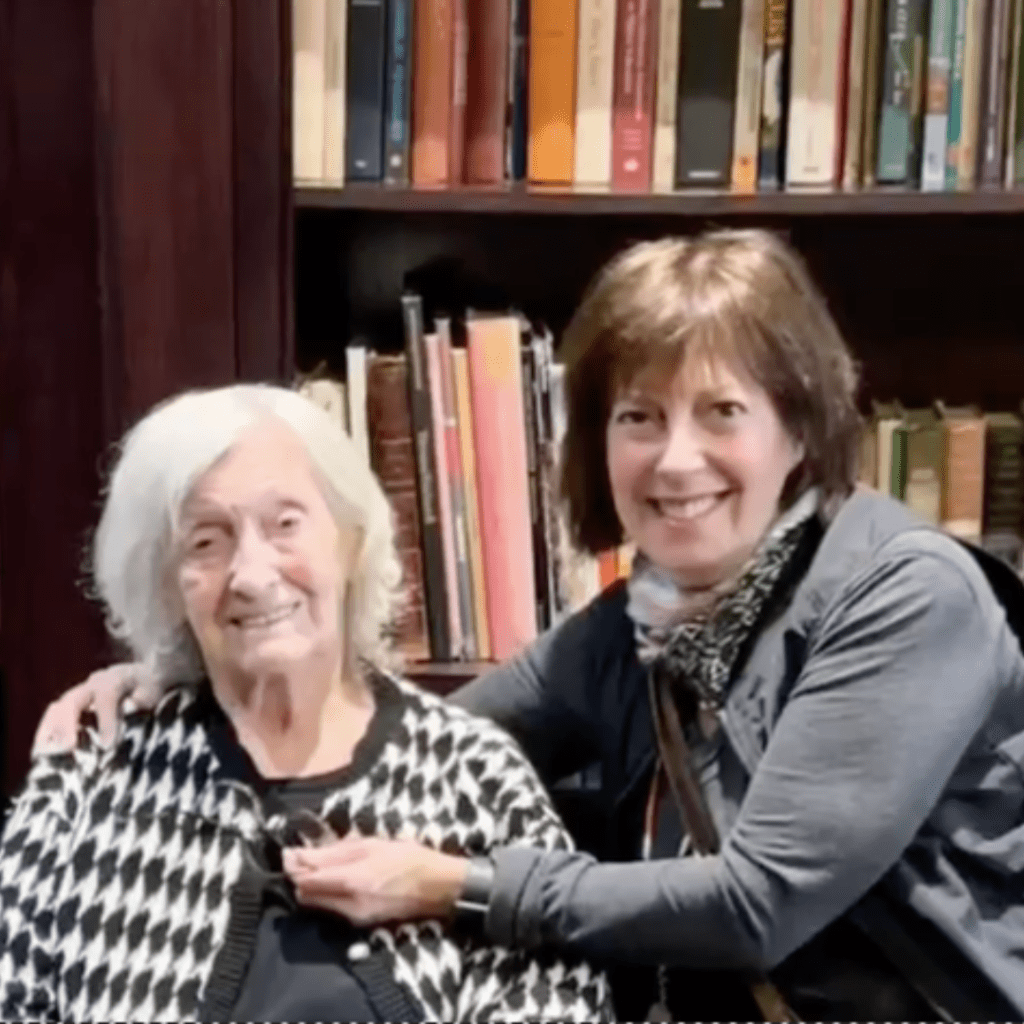 Grieving One of the Last Holocaust Survivors - photo of Hanka an older lady wearing a black and white cardigan seated in a chair and her daughter Debbie who is giving her a side hug, she is wearing a grey sweter. Both are smiling.